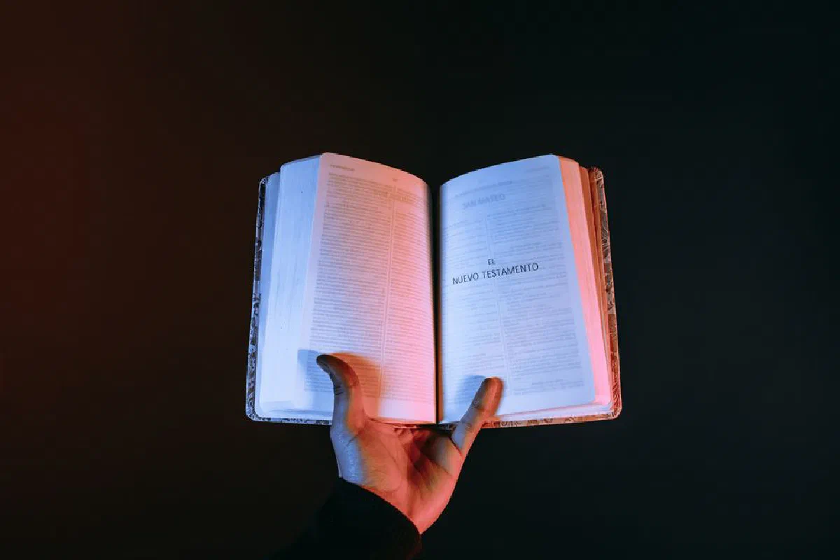A hand holds an open Bible displaying the New Testament against a dark background.