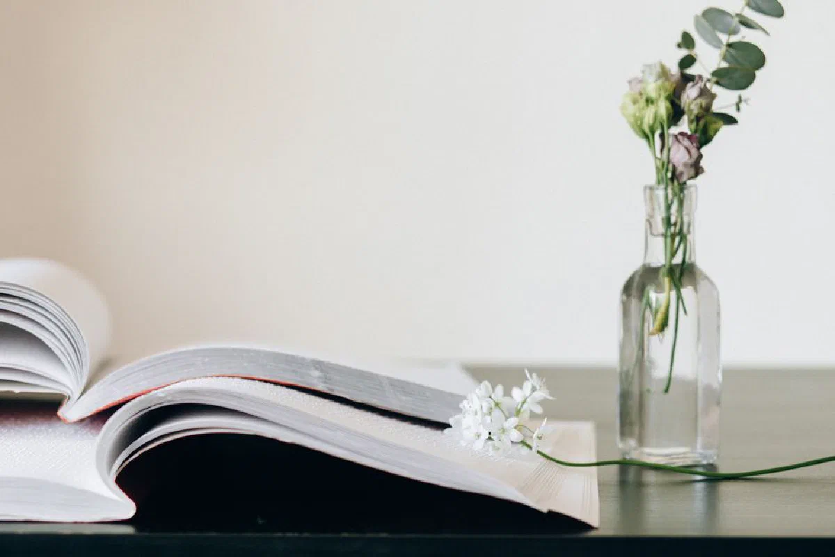 A serene still life featuring an open book next to a glass vase with delicate flowers on a dark surface.