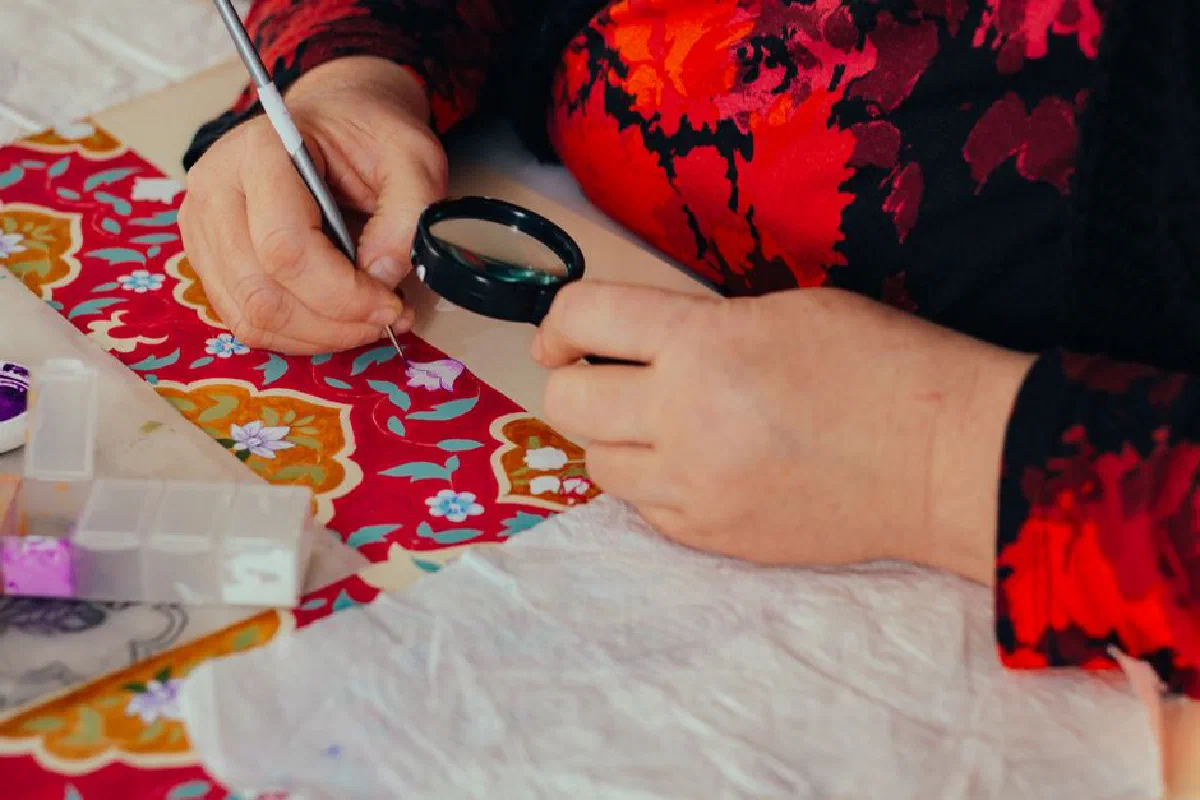 Close-up of a person painting intricate floral designs on fabric using magnifying glass.