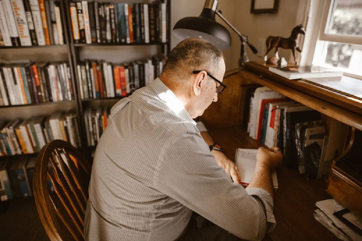 Adult man writing in a notebook at a wooden desk in a cozy home office library.
