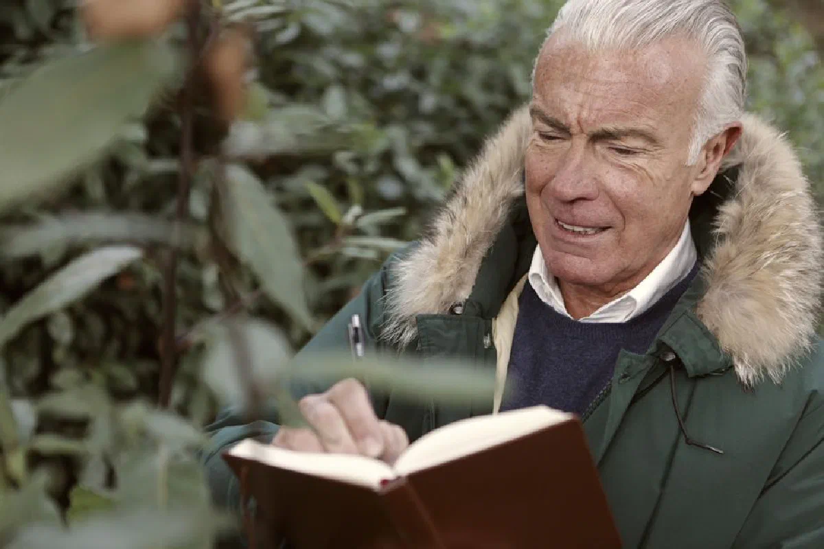 Senior man in a fur-lined coat writing in a notebook outdoors.