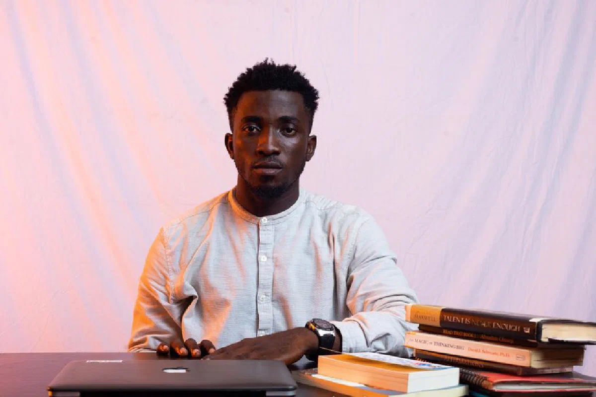 An African man sitting at a desk with a laptop and books, looking serious and focused indoors.