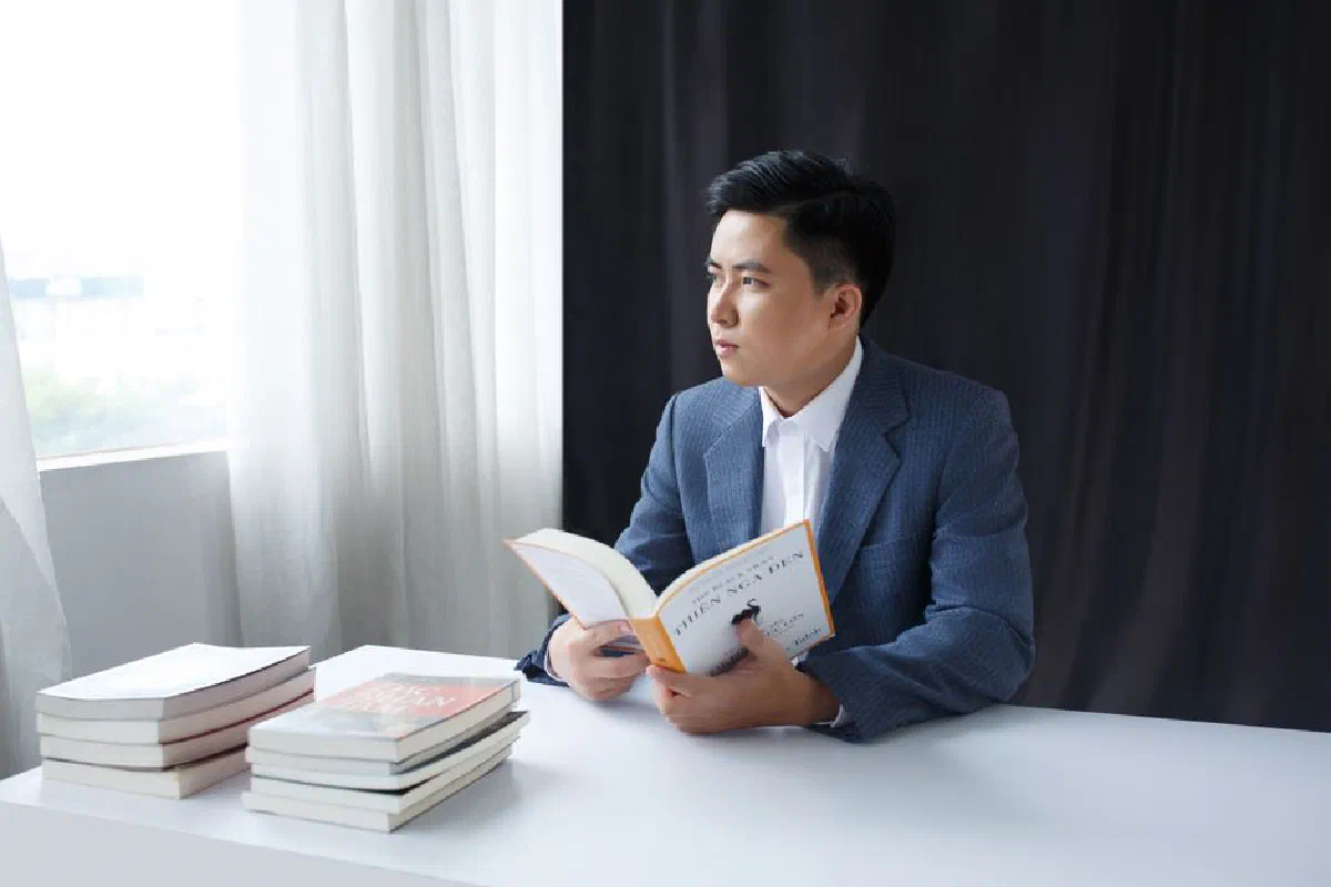 A young man in a suit thoughtfully reading a book at a table by the window.