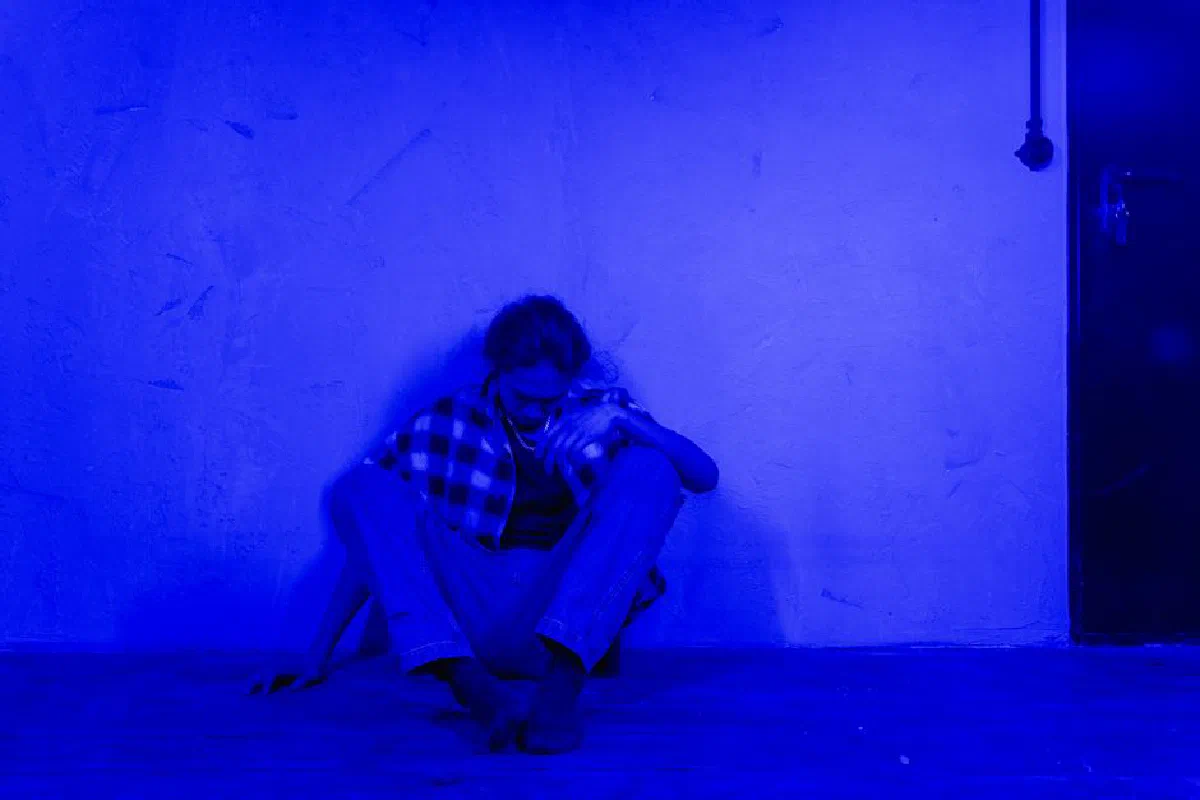 A woman sits alone indoors, illuminated by a blue light, expressing emotions of solitude and contemplation.