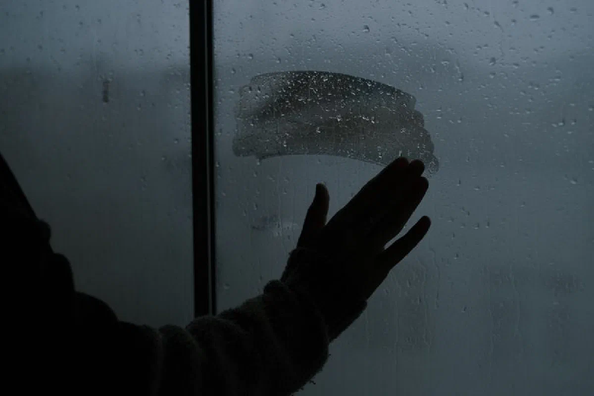 Silhouette of a hand touching a wet, misted window during rain, creating an emotional ambiance.