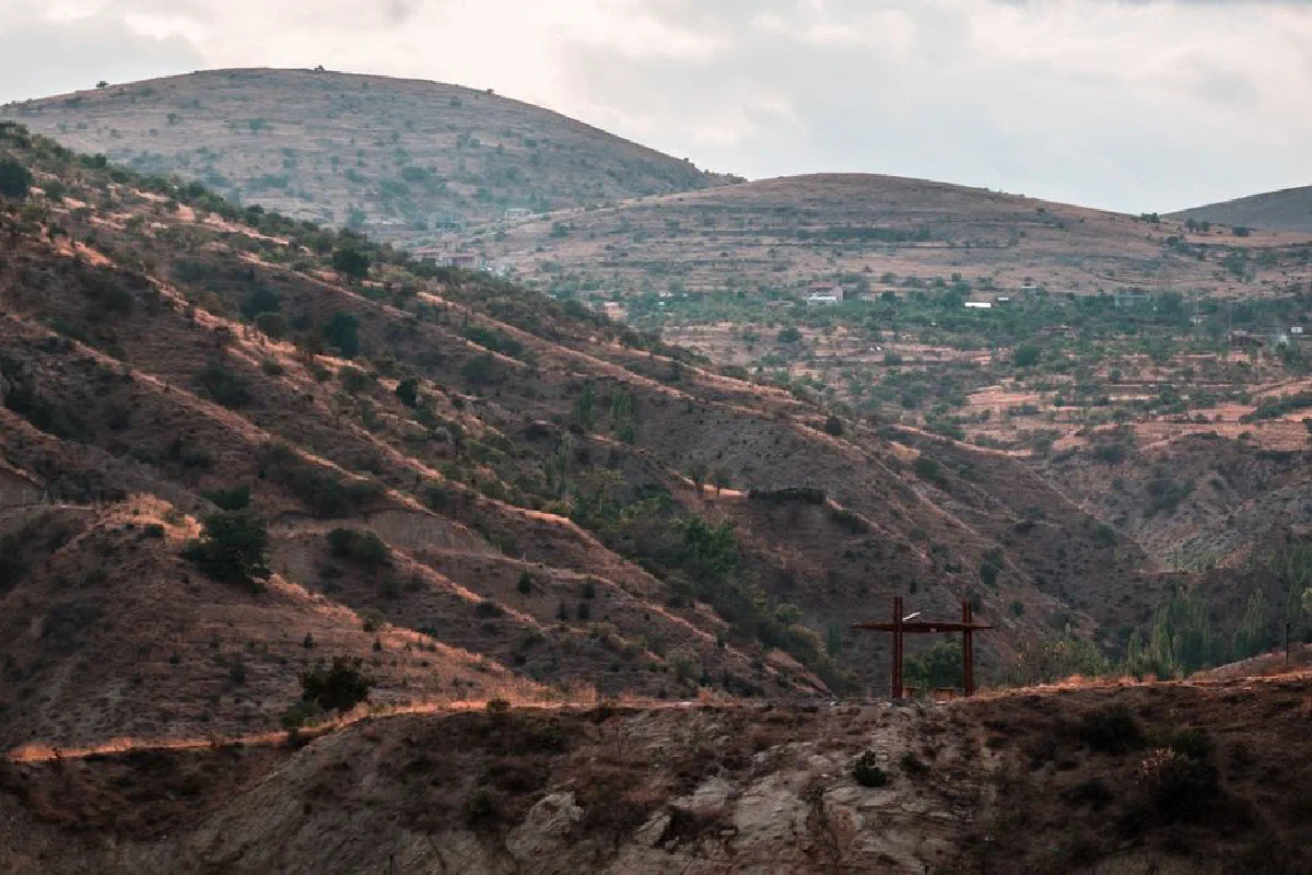 Desolate hills with sparse vegetation and a visible cross under cloudy skies.