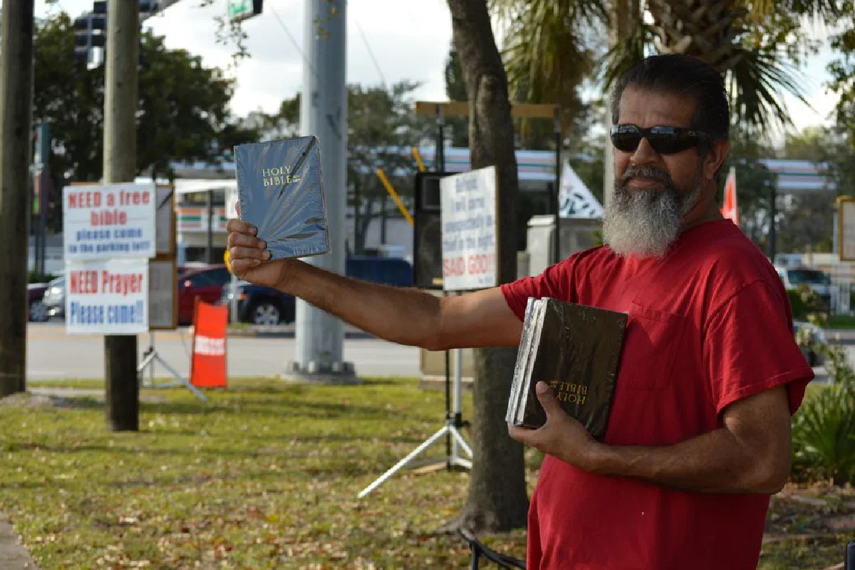 A man offers free Bibles on a sunny day, promoting Christianity and community outreach.