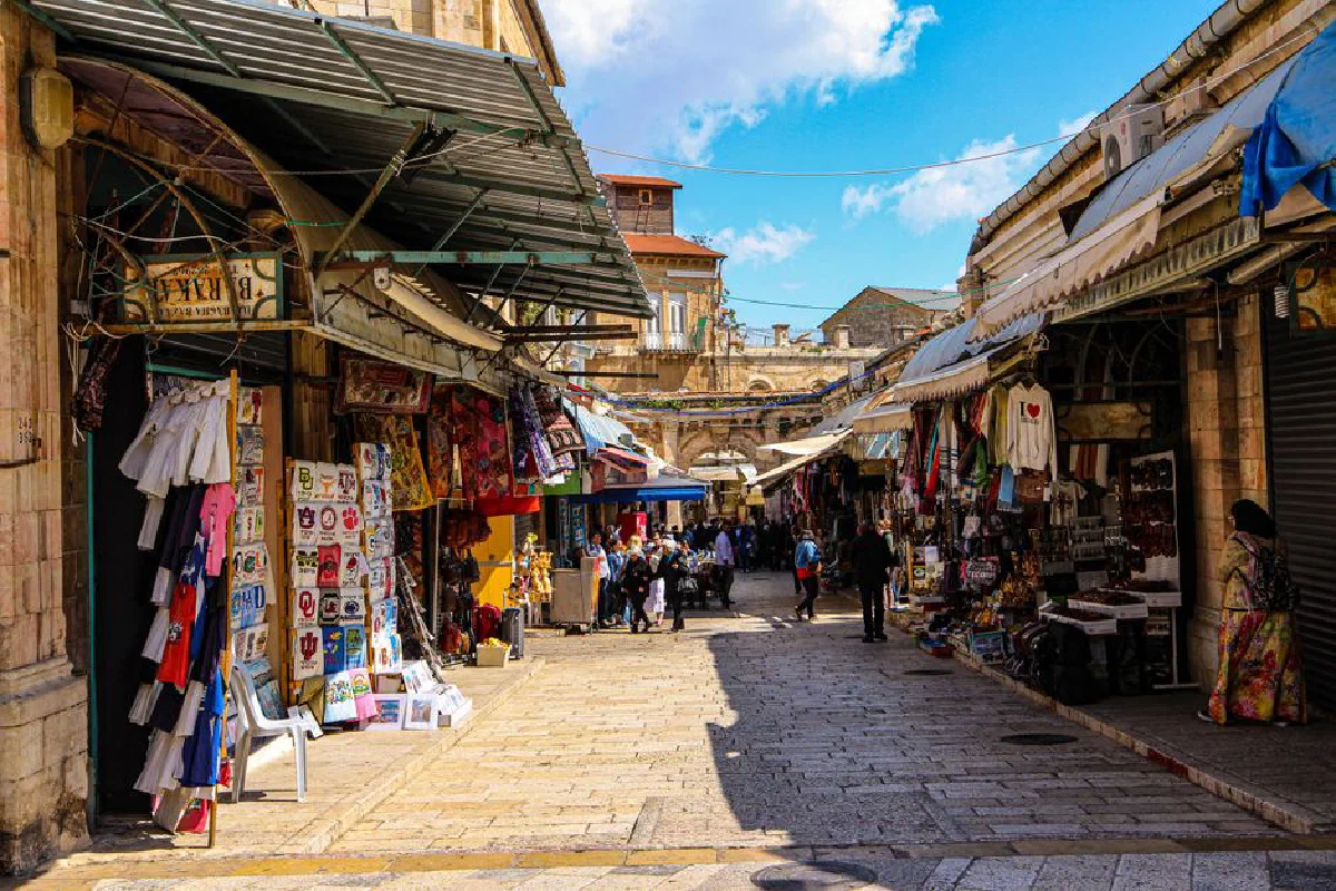 Bustling market alley in Jerusalem's Old City featuring shops and tourists under a sunny sky.