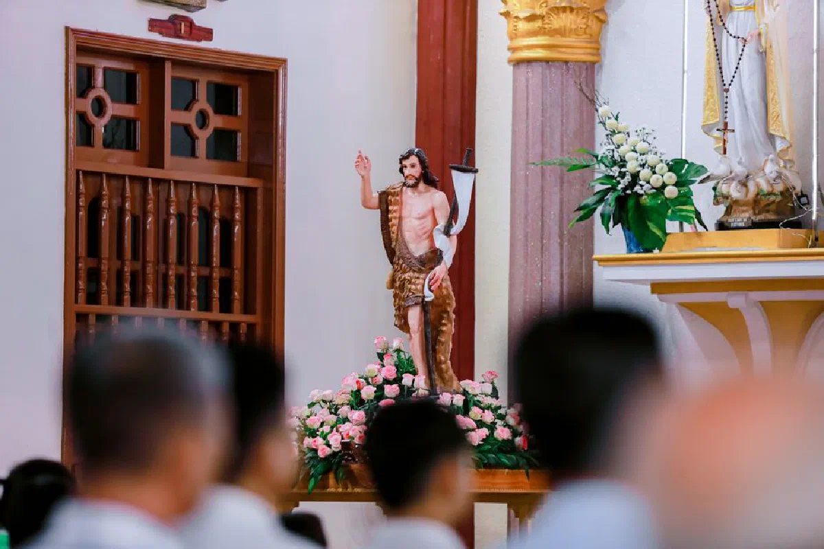 A religious statue surrounded by pink flowers inside a church during a gathering.