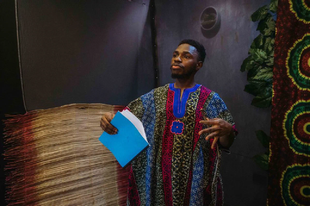 A man wearing vibrant traditional attire stands indoors while holding blue papers.