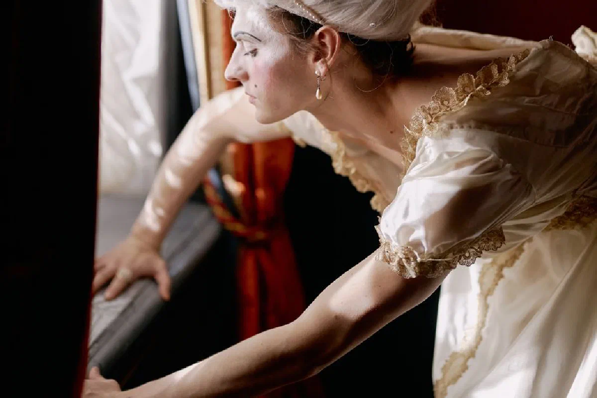 A performer in a vintage dress poses thoughtfully by a window, creating an artistic and dramatic atmosphere.