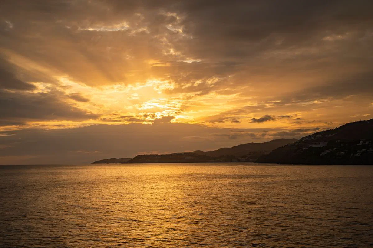 A breathtaking golden sunset casting radiant beams over the ocean horizon with silhouetted coastal hills.