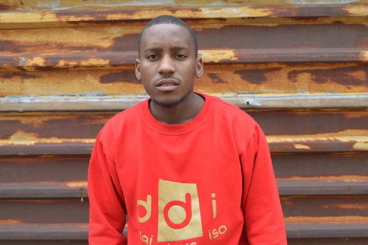 Casually dressed young man standing in front of a rusty wall wearing a red sweatshirt.