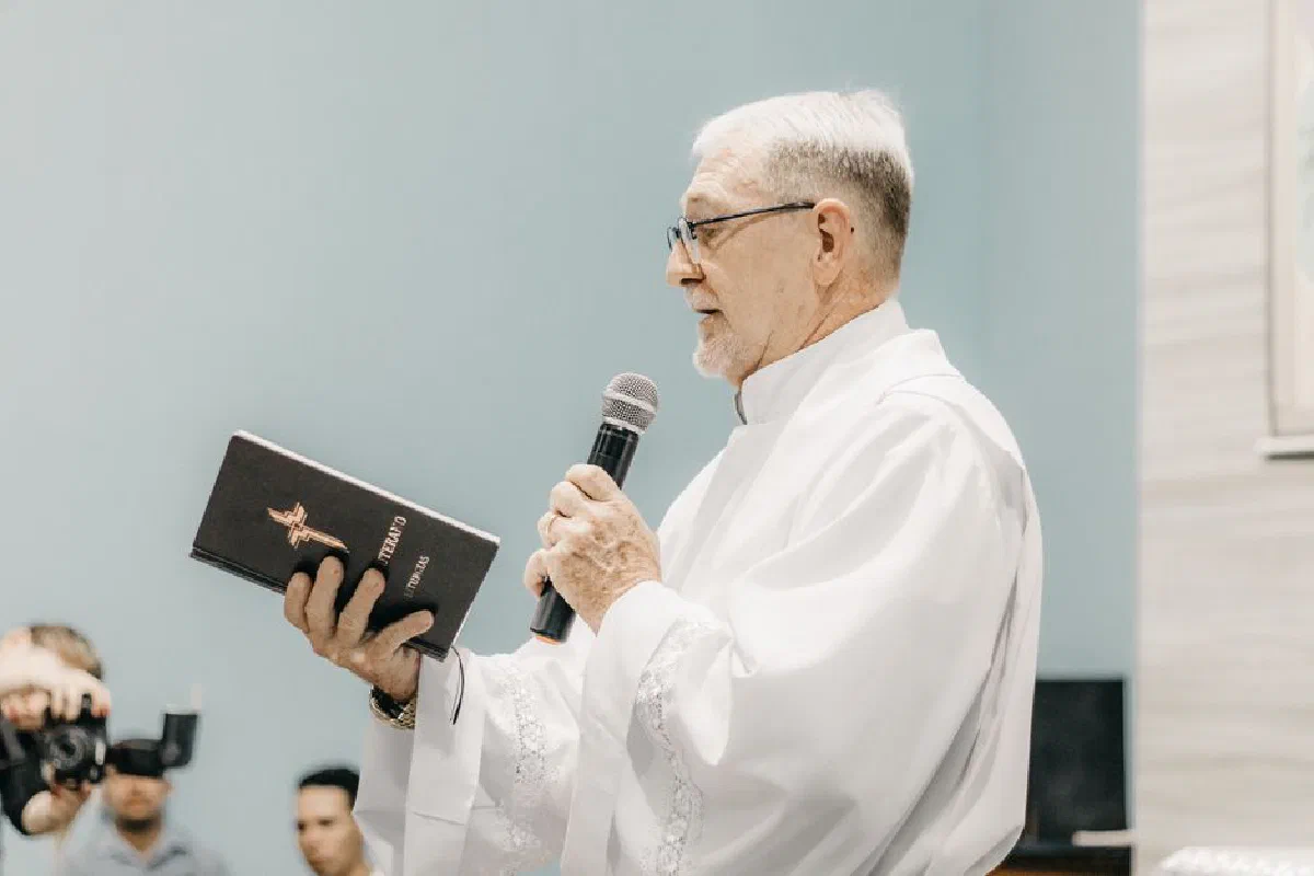 Priest in white robe holding Bible and microphone during church ceremony.