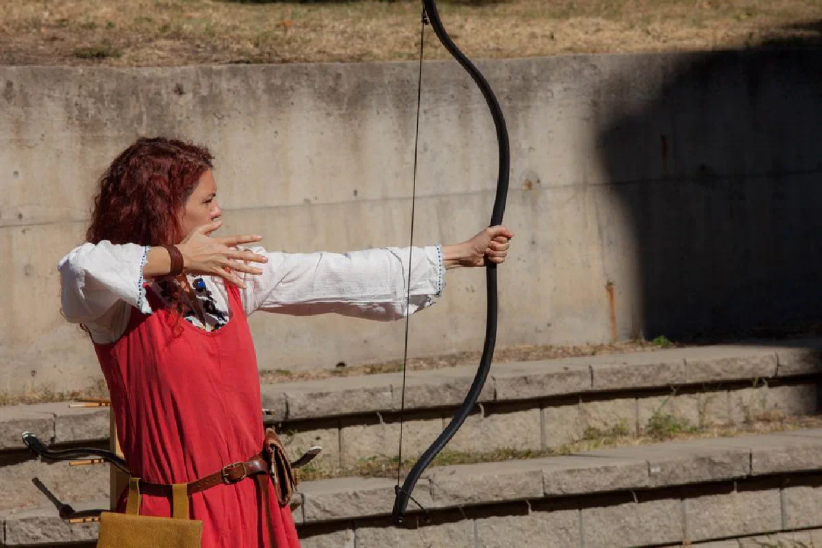 A woman in historical costume aiming a bow and arrow outdoors, demonstrating archery skills.