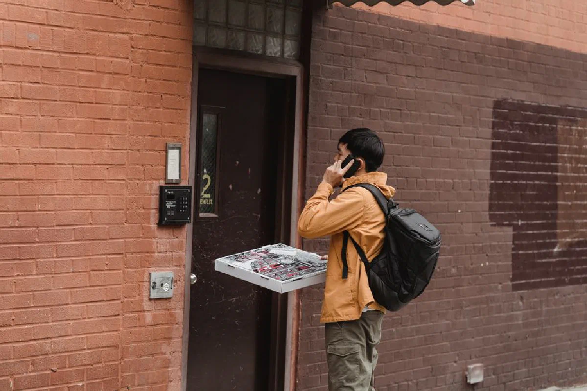 Man delivering pizzas, talking on cellphone, standing at apartment door with delivery bag.
