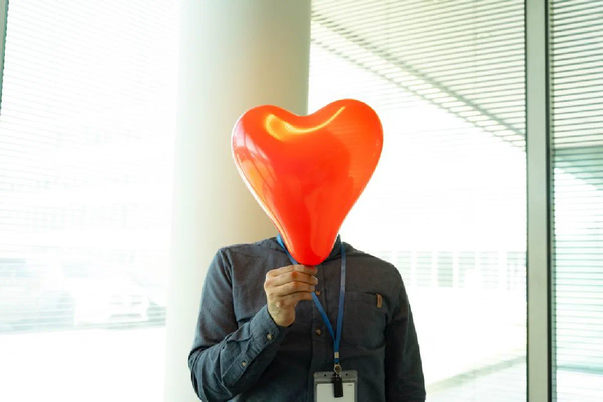 Humorous concept of a man holding a heart-shaped balloon over his face in an office setting.