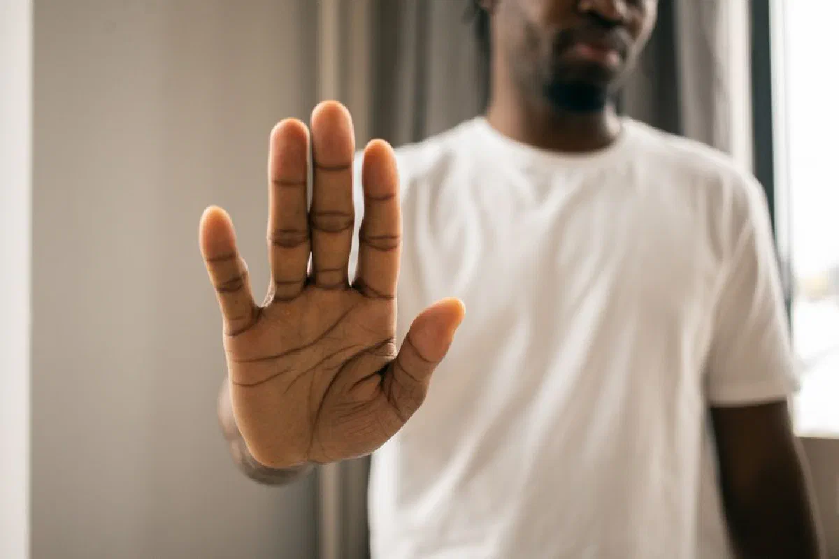 Crop anonymous African American male in casual white t shirt showing stop sign with hand while standing in light room