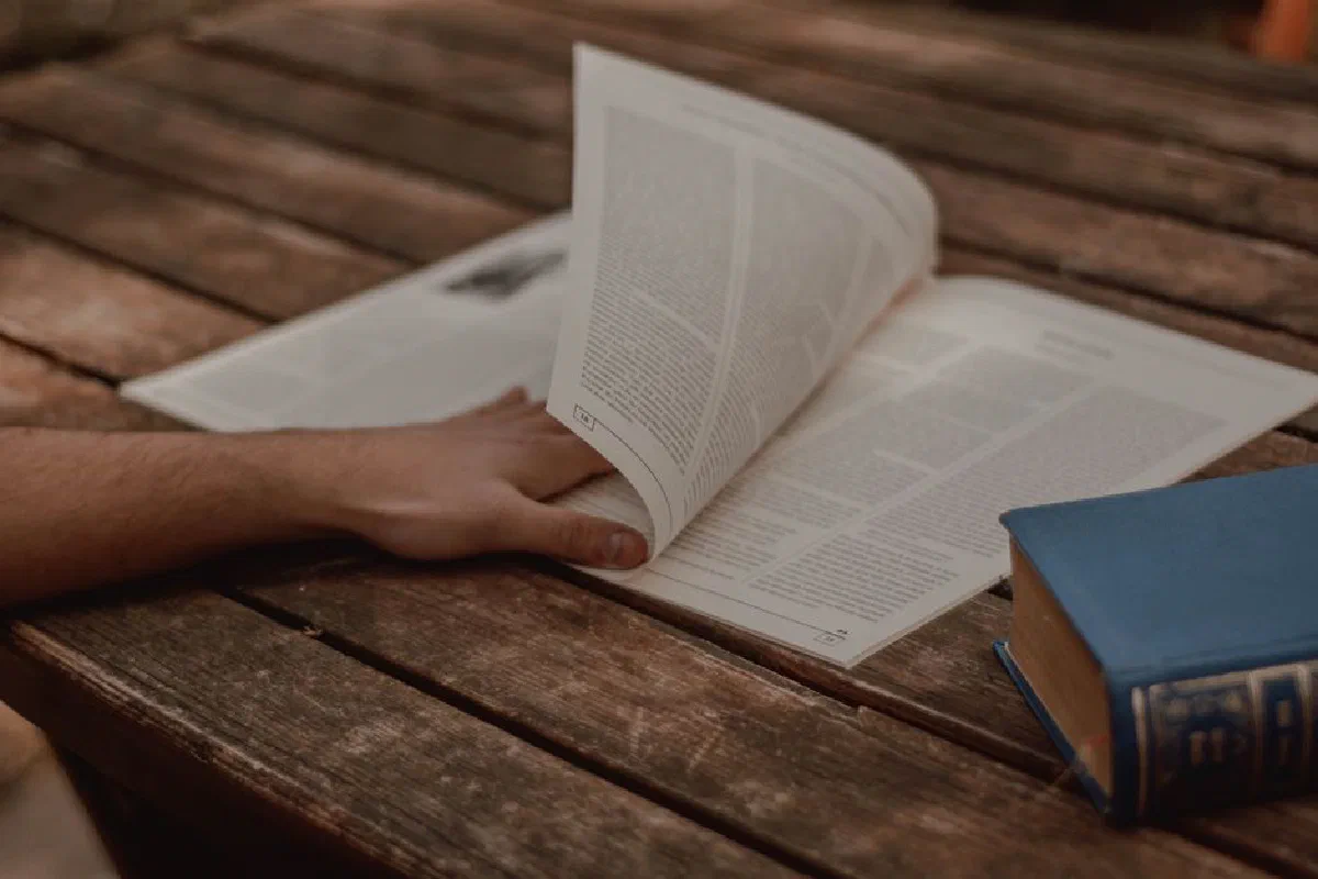 From above of crop unrecognizable male reading magazine with text in columns near old book