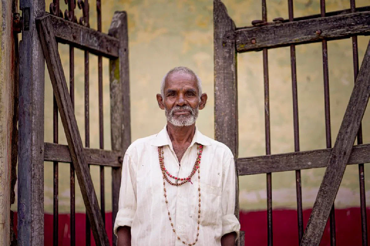 Portrait of an elderly South Asian man standing by a rustic gate in Melukote, India.