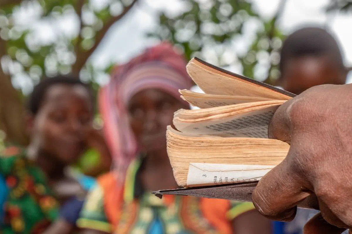 A close-up of a hand holding a book with blurred villagers in the background.
