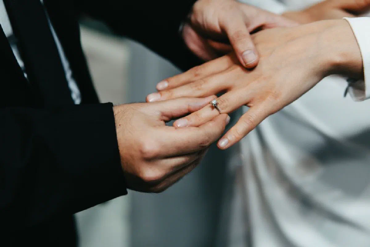 A groom places a wedding ring on the bride's finger during a close-up ceremony shot.