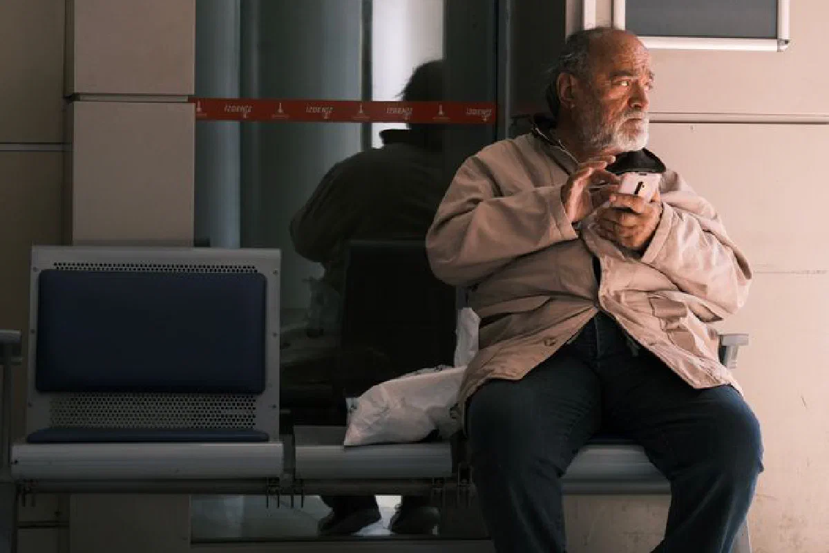 Elderly man holding a coffee cup, sitting alone on a bench indoors.