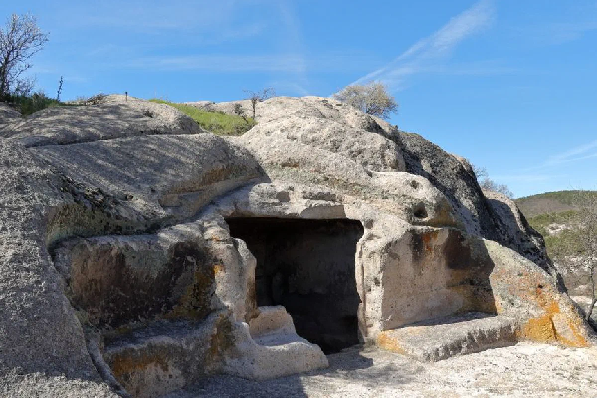 Explore the entrance of a historic rock cave under a bright blue sky, with distant hills in the background.
