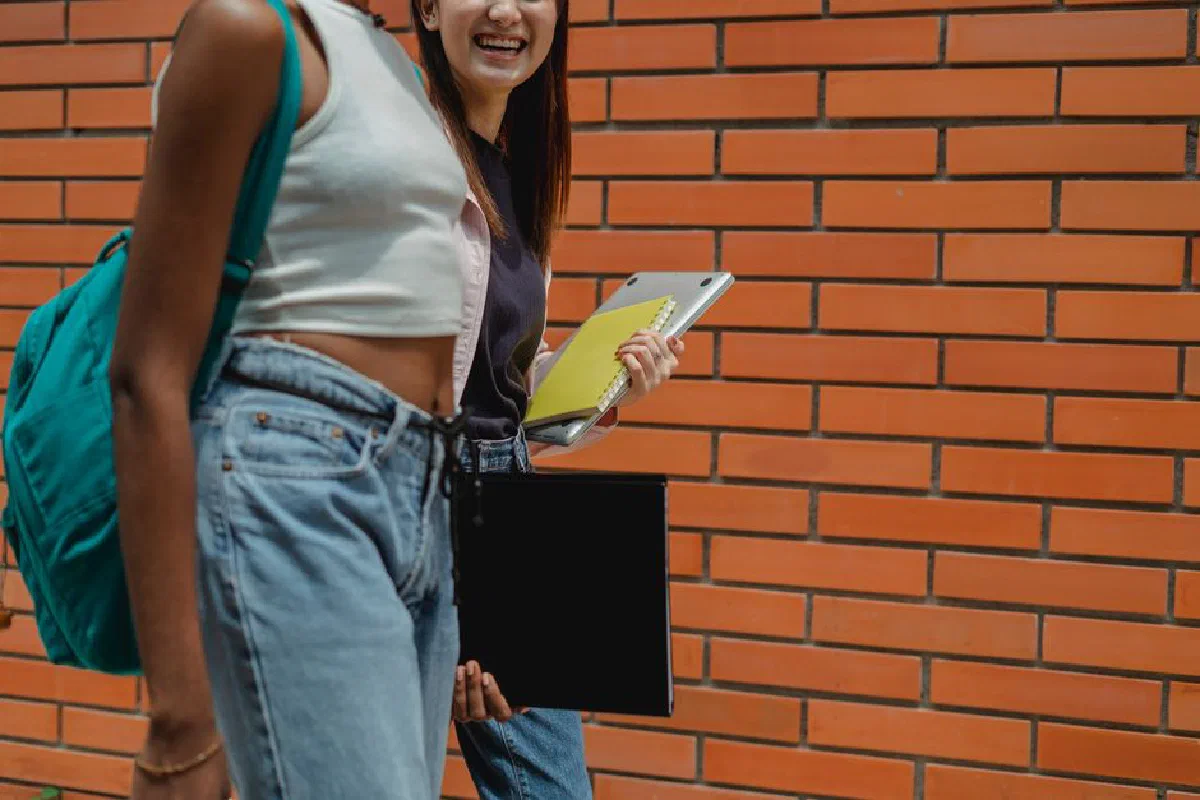 Side view of crop anonymous female friends in casual wear with laptop and copybooks strolling near brick wall and speaking to each other
