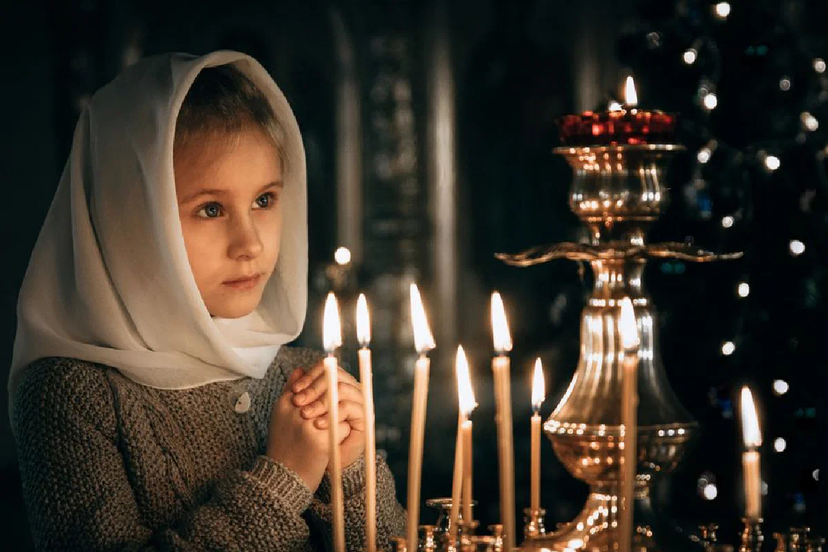 A young girl in a headscarf prays solemnly in candlelight, creating a serene atmosphere.