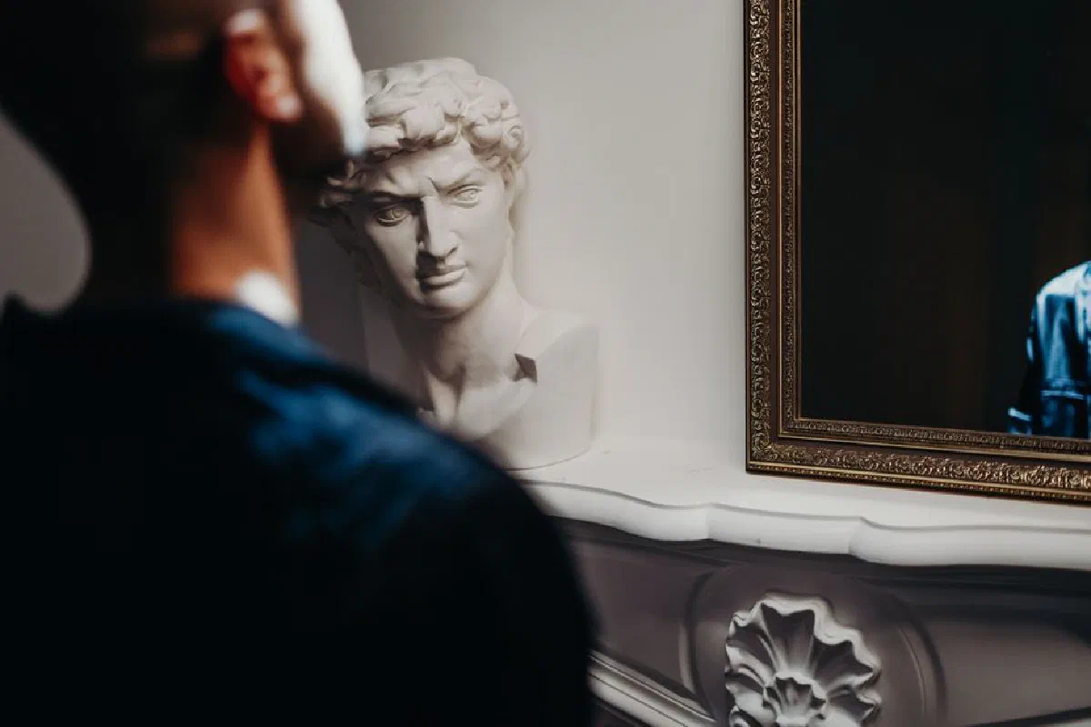 A man gazes into a mirror beside a classic sculpture in a sophisticated room.
