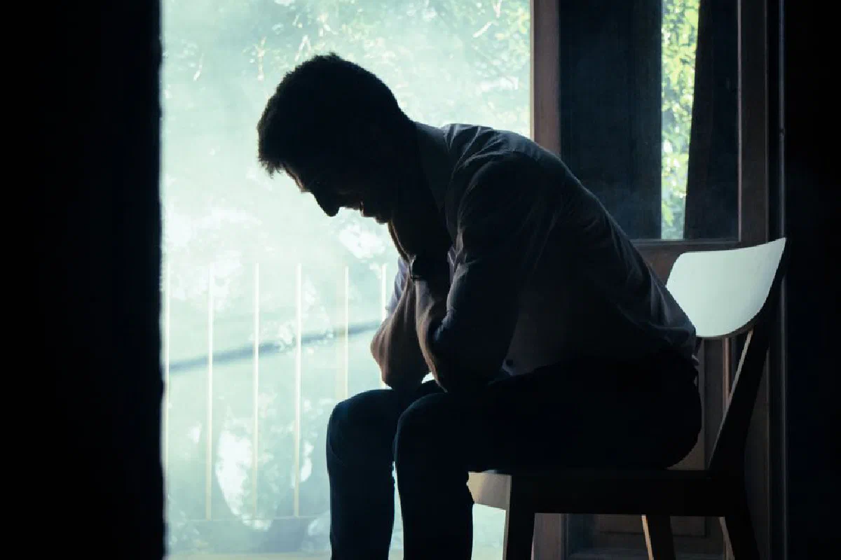 A man sits alone in a moody indoor setting, silhouetted against a bright window, deep in thought.