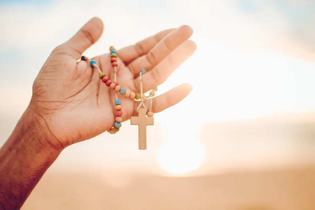 A hand holding a colorful rosary with a wooden cross against a bright sunrise background.
