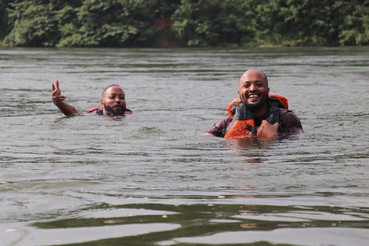 Two men with life jackets smiling while swimming in a serene river environment.