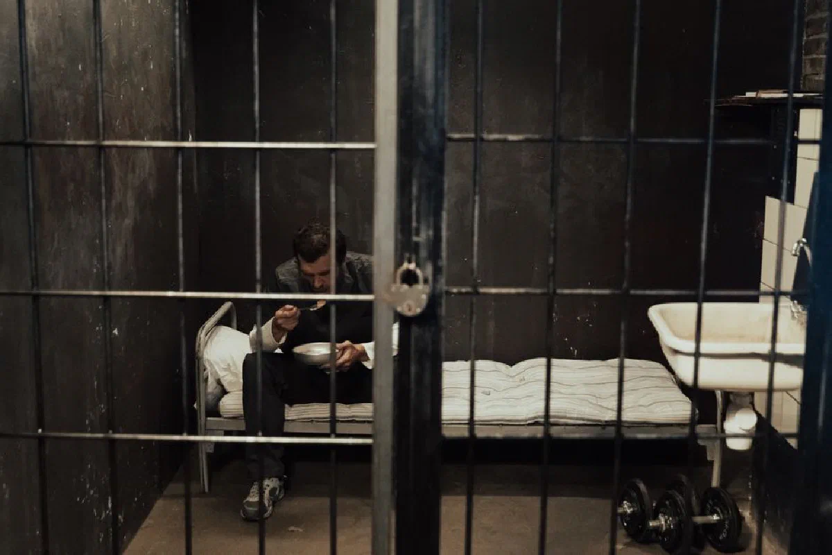 A prisoner sits alone in a dark cell, eating with focus in a moody environment.