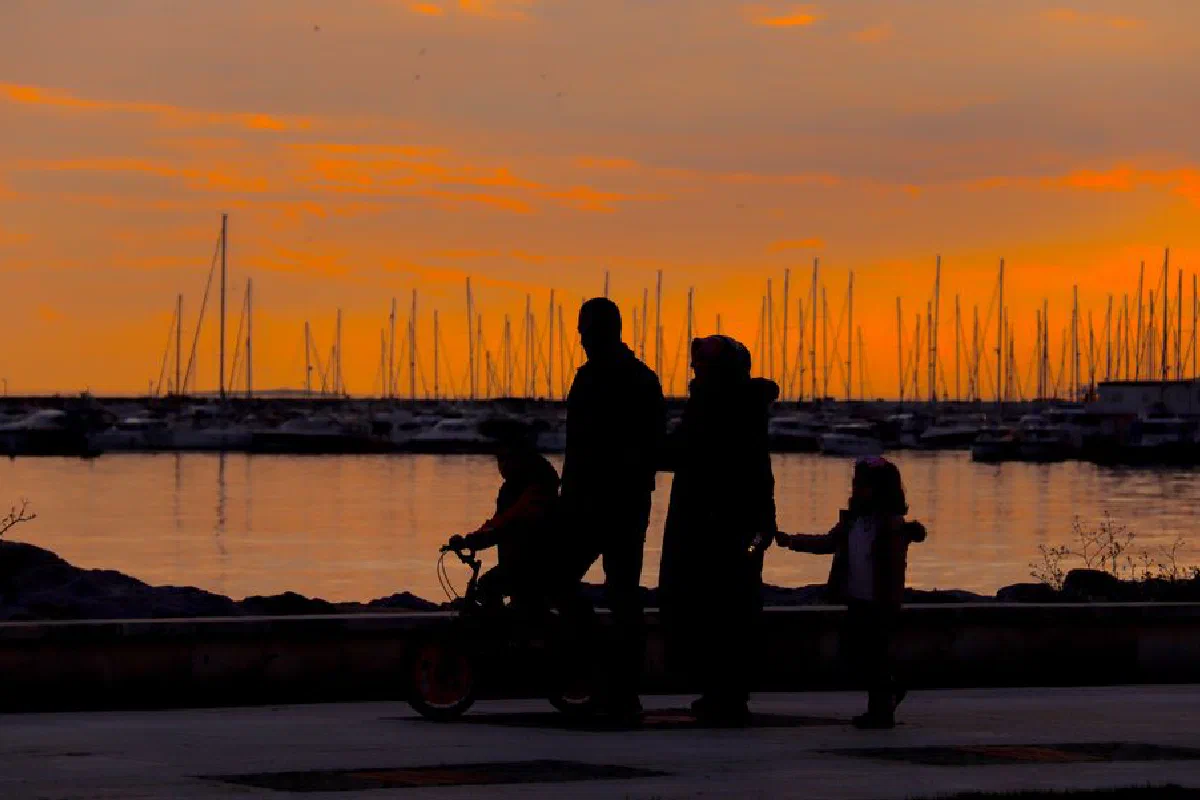 A silhouette of a family walking by the harbor at sunset with boats in the background.